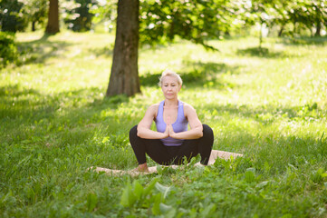 Naklejka na ściany i meble Middle-aged woman doing yoga in sportswear outdoors in a city park in Namaskarasana - Seated Prayer Pose. Concept of stretching, pilates, healthy lifestyle.
