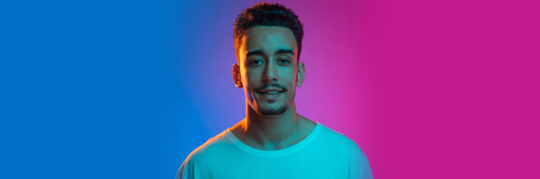 Positivity. Young man in casual white T-shirt posing, looking at camera over gradient blue pink background in neon light. Banner, flyer. Concept of emotions, facial expression, sales, ad, fashion