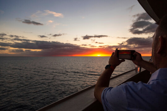 Man taking a smartphone photo of a beautiful sunset while enjoying an evening on a luxury cruise ship. Cruise vacations are relaxing and enjoyable