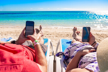An adult couple relaxing on the beach looking at their cell phones as they enjoy their vacation resting in beach chairs on a beautiful sunny day