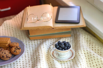 Plate of cookies, cup of tea, fresh blueberries, dry oranges, stack of books, reading glasses and tablet on the table. Hygge at home. Selective focus.