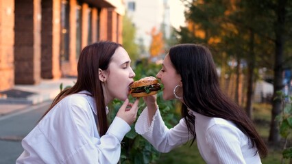 Two teenage friends biting the same hamburger at the same time, two friends eating junk food on the...