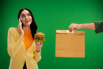 smiling woman holding burger and talking on smartphone near courier with paper bag isolated on green.