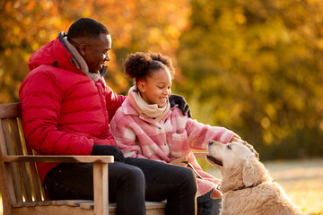Family Shot With Father And Daughter Sitting On Bench With Pet Dog On Walk In Autumn Countryside