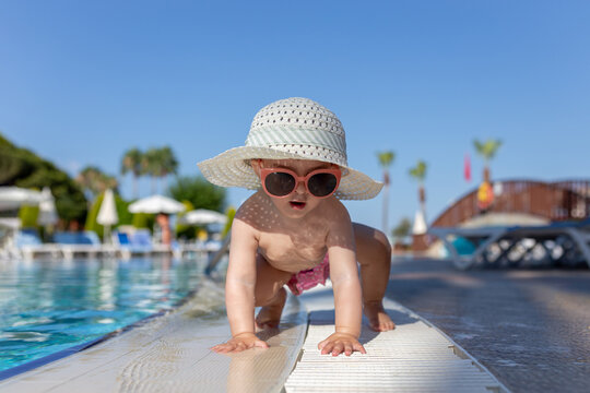 Baby girl wearing sun hat and sunglasses while crawling next to the swimming pool outdoors.