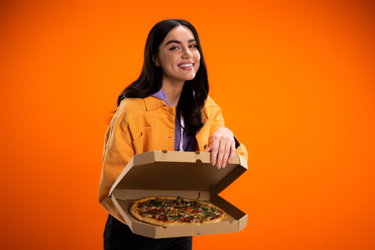 smiling brunette woman showing tasty pizza in carton box while looking at camera isolated on orange.
