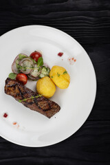Grilled beef skirt steak meat with potato and vegetables on white plate on wooden black background