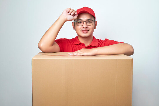 Friendly- ooking asian delivery guy in red cap and uniform, courier service lean on cardboard box or client package, saluring as making order