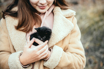 Smiling caucasian teen girl with long loose hair wearing faux fur coat and holding black small homeless kitten in her hands outdoors in autumn day. Taking care and protecting