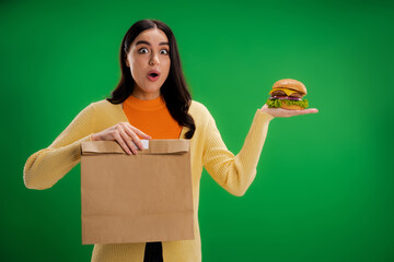 amazed woman with tasty burger and paper bag looking at camera isolated on green.