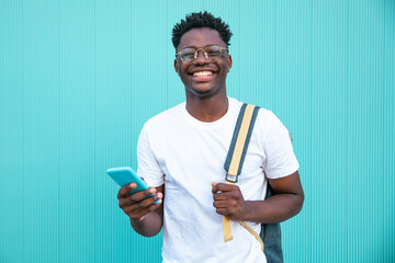 Portrait of happy young man smiling with a mobile phone and a backpack on turquoise background -...