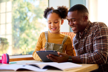 Father At Home Helping Daughter In Lounge As She Does Homework Using Digital Tablet