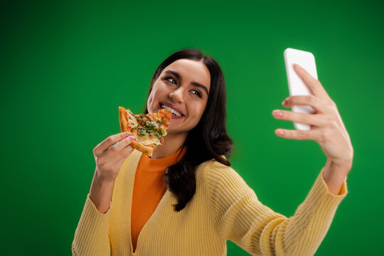 cheerful woman biting piece of pizza and taking selfie on smartphone isolated on green.