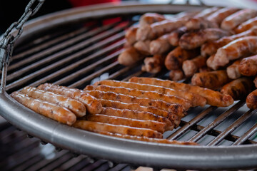 Process of grilling fresh meat sausages on big round hanging grill at summer local food market: close up view. Outdoor cooking, barbecue, gastronomy, cookery, street food concept
