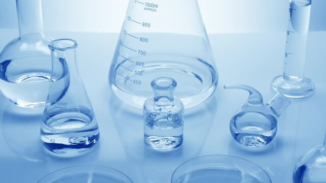 Close-up crane shot of water drops drip into reagent bottle surrounded by lab glassware with water on pale blue background | Background shot for skin moisturizing cosmetics commercial