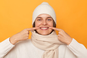 Smiling happy cheerful young caucasian woman wearing white sweater, hat and scarf, pointing at her teeth, showing result after whitening or visiting dentist, posing isolated over yellow background.