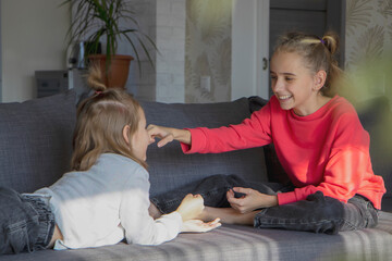Two little sister girls play at home in the living room, look at each other, laugh and smile. The concept of warm relationships, love, trust and understanding in the family.