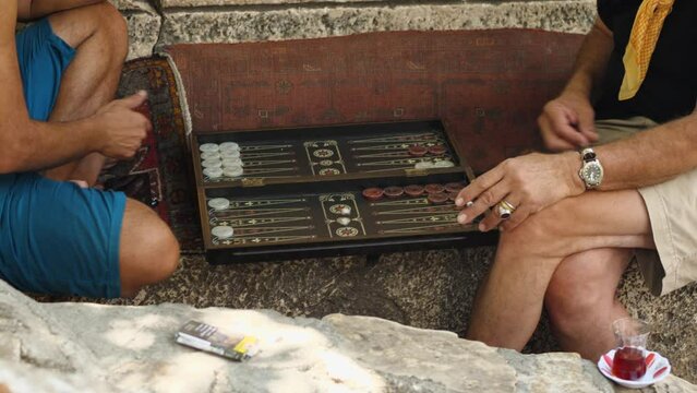 Close-up of two men playing backgammon on the street in Turkey. One man is smoking. A cup of tea is next to him. Backgammon for the street