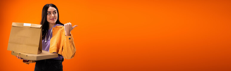 smiling woman with food packages pointing with thumb and looking away isolated on orange, banner.
