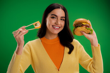 cheerful woman holding tasty burger and fresh sushi while looking at camera isolated on green.