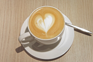White Cup of Coffee with Milk Foam on Wooden Table in a Cafe. Cup of Hot Latte and Saucer with Spoon. Delicious Stylish Cappuccino Art Coffee. Closeup of Heart Shape Drawing in Foam. Coffee Shop.
