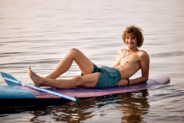 Fototapeta premium Relaxed caucasian male surfer lying on surfboard after surf on high waves. Sporty young smiling man enjoy the rest alone, looking at camera. Extreme sports, youth active lifestyle concept