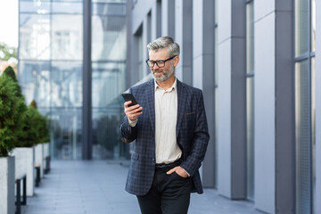 Successful gray-haired businessman walks outside office building, mature boss in glasses uses phone, man smiles and types message, reads online news and browses internet pages.