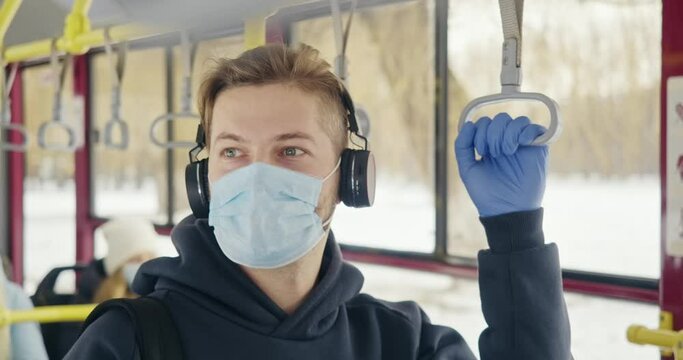 bFront view of oy with earphones, wearing medical mask and gloves. Passenger standing on bus, listening to music in earphones, looking around. Concept of public transport.