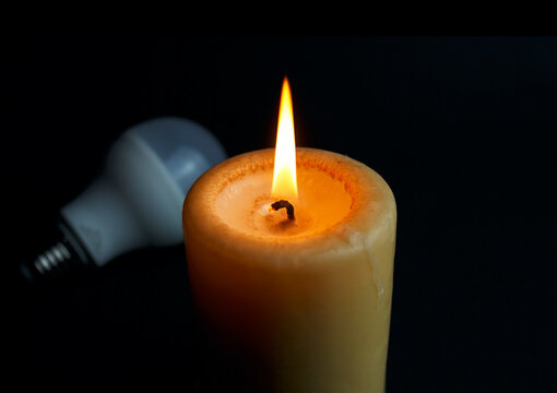 Burning candle and switched off light bulb in darkness. Blackout, electricity off, energy crisis or power outage, concept image.