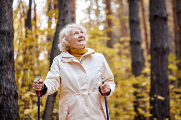 Senior aged woman standing with nordic walking poles in autumn park. Healthy lifestyle concept. Mature caucasian female resting after exercise outdoors, laughing smiling, enjoying time outdoors