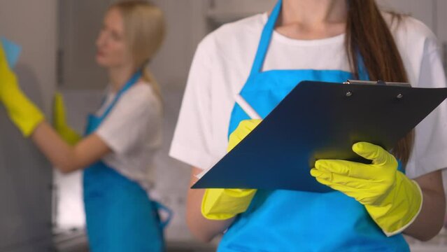 Young woman of professional cleaning company in uniform marks and writes down in tablet list of work done to clean room. In background they clean wash . Check and control with recording and fixation.