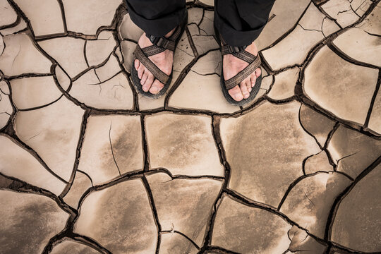 Feet in sandals standing on cracked mud.