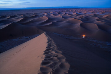 A campfire glows at twilight besides a tent among the sand dunes of Erg Zehar.