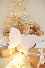 little blond boy near a wooden Christmas tree with craft gifts. A boy with angel wings and New Year's decor. Christmas photo zone. New Year's gifts. To believe in the Christmas miracle