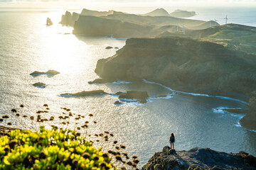 Backpacker woman enjoys panoramic view from a steep cliff over the seascape and along the rugged foothills of Madeira coast at sunrise. Ponta do Bode, Madeira Island, Portugal, Europe.