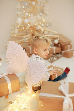 little blond boy near a wooden Christmas tree with craft gifts. A boy with angel wings and New Year's decor. Christmas photo zone. New Year's gifts. To believe in the Christmas miracle