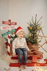 Obraz na płótnie Canvas a little boy sits on a sled near a Christmas tree. Christmas children's photo session in the studio. a child in a cap of Santa Claus. New Year's mood. New Year's photo zone in a photo studio. 