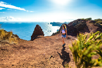 Backpacker woman enjoys hike along a steep cliff overlooking the sea and the rugged foothills of Madeira's coast in the morning. Ponta do Bode, Madeira Island, Portugal, Europe.