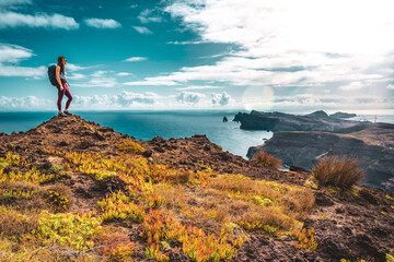 Obraz premium Backpacker woman enjoys panoramic view from a steep cliff overlooking the sea and the rugged foothills of Madeira's coast in the morning. Ponta do Bode, Madeira Island, Portugal, Europe.