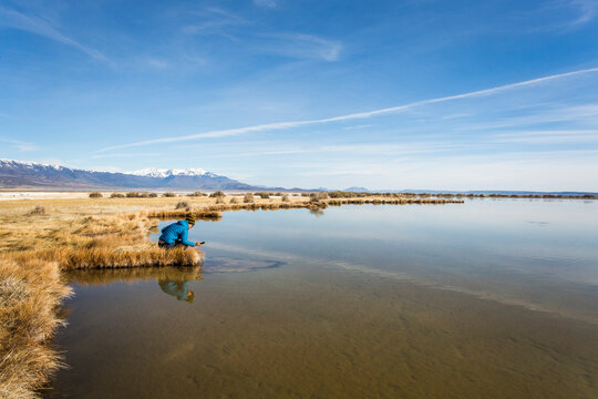 BORAX LAKE, HARNEY COUNTY, OR, USA. A young woman in a blue coat crouches to look into a shallow hot spring pond in a vast, dry landscape.