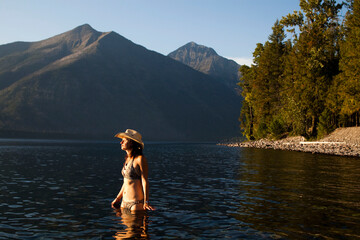 A young woman, wearing a cowboy hat, enjoys the calm, blue waters of a lake as the sun sets near mountains in the distance.
