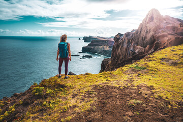 Obraz premium Backpacker woman enjoys panoramic view from a steep cliff overlooking the sea and the rugged foothills of Madeira's coast in the morning. Ponta do Bode, Madeira Island, Portugal, Europe.