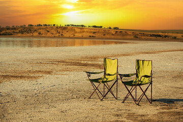 Two tourist chairs in a desert area on a lake at sunset. Secluded vacation in the wild.