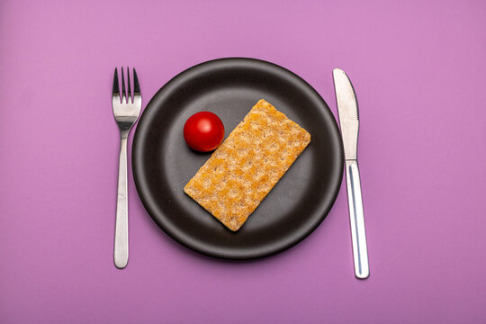 A single tomato and a slice of crispbread on a plain black plate and cutlery with signs of use against a purple background