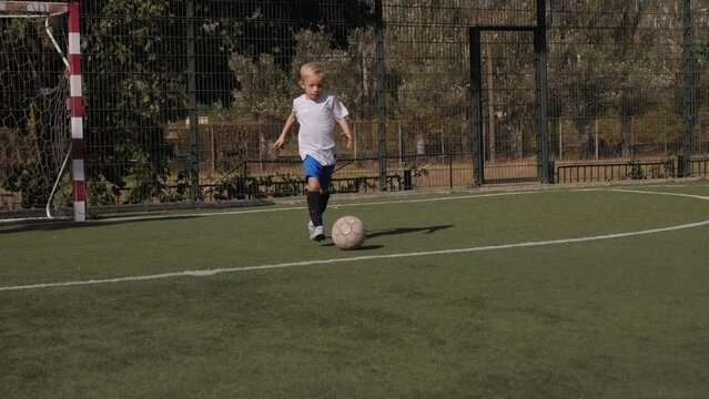Little boy in a football uniform trains on the football field, practicing dribbling and feints in sunny weather.