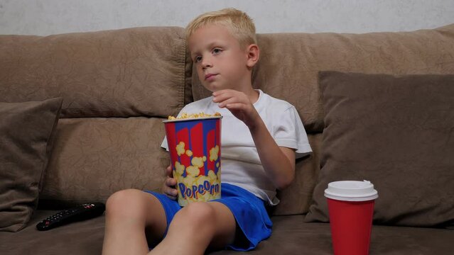 A little boy is watching a football match on TV, sitting at home on the couch with popcorn.