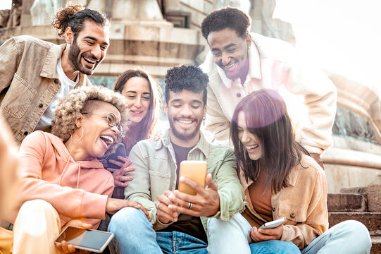 Multiethnic group of smiling people using mobile phone outdoors -  Cheerful guys and girls friends having fun watching at digital display sitting in the city street - Social media lifestyle.