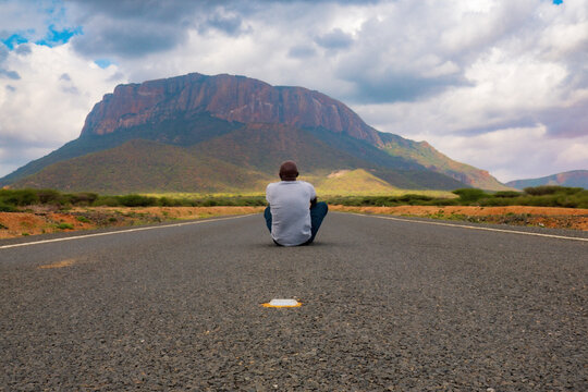 man sitting on the an empty highway against the background of Mount Ololokwe in Marsabit County, Kenya