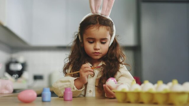 Little girl painting eggs with pink paint for a traditional spring dinner. Happy Easter. Kid making easter decoration at home.Cute child wearing bunny rabbit ears on easter day preparing for holiday
