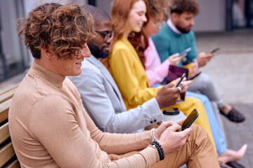 Curly young pleasant male sit surfing the Net during break outdoors, sitting with mixed race colleagues. Caucasian handsome guy in brown shirt use modern smartphone, have rest in city downtown